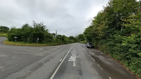 A rural road junction with hedges either side on a cloudy day. There is a small layby on the right and a turning on the left.