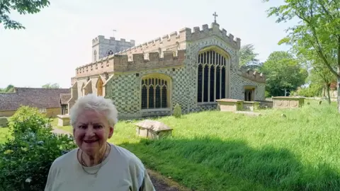 Ewelme historian Carol Sawbridge smiling for a picture in front of Ewelme Church. It is a sunny day.