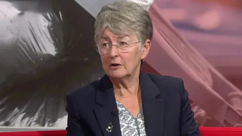 BBC A woman with short grey hair, sitting on a red sofa in a television studio. She is wearing a navy blazer and blue and white top. 