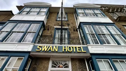 Martin Giles/BBC A picture of of the outside of the Swan Hotel. It has three stories of square sash windows which are have a blue facade. "Swan Hotel" is written in gold lettering. 