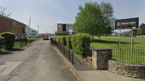 The exterior of Southmoor Academy and its entrance road. There is a sign on a land of green grass to the right and then a single track road which has a 5mph sign and a barrier which is up. There is a red Post Office car which is travelling into the car park. The main school building is in the distance and a two-storey house is on the left.