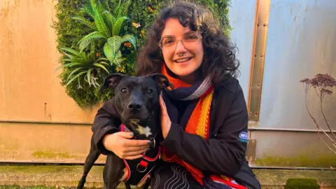 Bristol Animal Rescue Centre A young woman with curly dark hair, a bright scarf and dark coat, smiles at the camera as she hugs a black dog, which is a type of staffordshire bull terrier cross