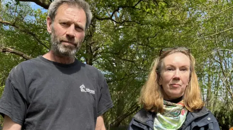 A man and a woman stood side-by-side. The man has a white beard and hair. The woman has blonde hair.