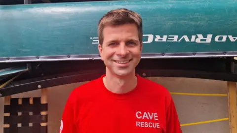 Devon Cave Rescue Organisation A man with dark hair, Olly Rees, in a red t-shirt that says 'cave rescue' on it