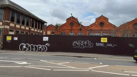 Oliver Conopo/BBC Red-brick facades with windows showing above the brown line of hoardings can be seen from the road. The corner of the transport office is on the left.