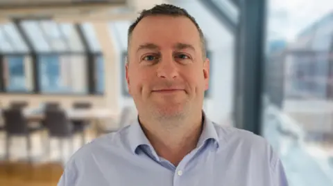 Pracedo Philip Young is standing in an office wearing a blue shirt and smiling.