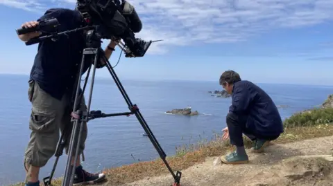 A man is crouched on a cliffside while another man films him with a camera mounted on a tripod. 