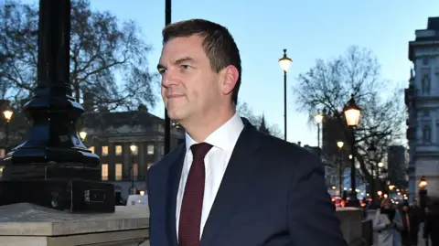 Olly Robbins walking down Whitehall in central London wearing a navy suit and a red tie