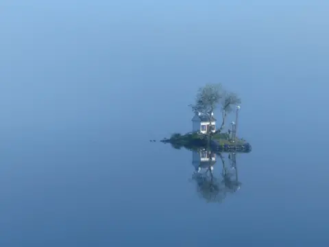 Rolande Berninger Blue sky and water fading into each other with a small island with a small white house and tree