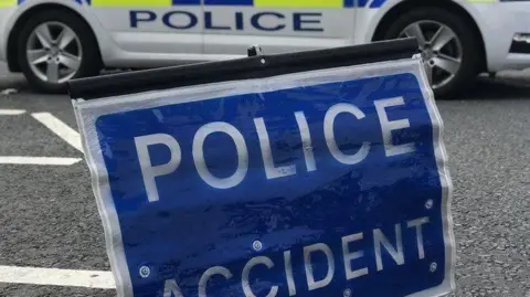 A blue rectangular sign with white lettering saying "police accident" resting on a road. Behind it is the lower part of the side of a police car in yellow, blue and white livery.