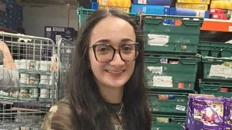 Joshua Holmes-Bright/BBC Davina Chipperlow is standing in front of green pallets of food. She has long dark hair and is wearing glasses and smiling.