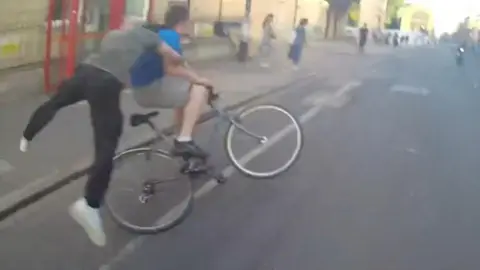 A man wearing a green t-shirt is tackling a man in a blue t-shirt whilst he is riding a bike 