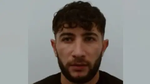 a police mugshot of a man with short black hair and a black beard who is wearing a black t-shirt