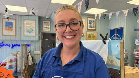 Sofie Bell wearing a blue polo shirt and company apron smiling at the camera with glasses on and her hair tied back with her ice cream shop in the background