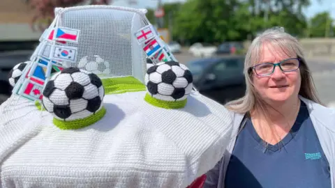 Chrissie Perry A woman with long blonde hair, fringe and glasses stands next to a post box topper with knitted white base, black and white footballs on green stands, a goal and tiny flags depicting national flags.