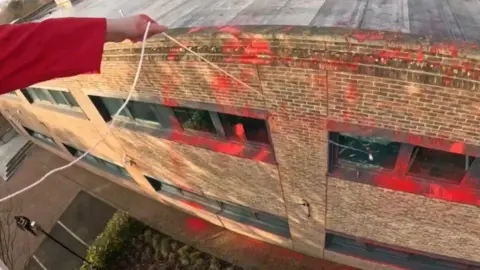 An extended arm, wearing a red long sleeve, swings a hammer on a rope through a broken window. Red paint covers the side of the building.  