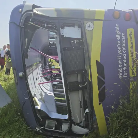 Jacko Thomas The front of a bus that has crashed and is overturned in a grassy field.