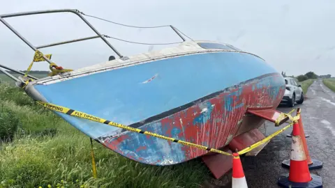John Devine/BBC A cabin cruiser boat, about 6m (20ft) long and 2m (6ft) wide lays on its side next to farmland, a narrow road can be seen in the background and more fields and trees. A silver car can be seen in front of the vessel.