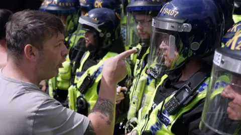 Getty Images A man with short head and tattoos points his finger in the faced of a police officer. The man appears to be saying things to the officer, who appears to be looking away from him.