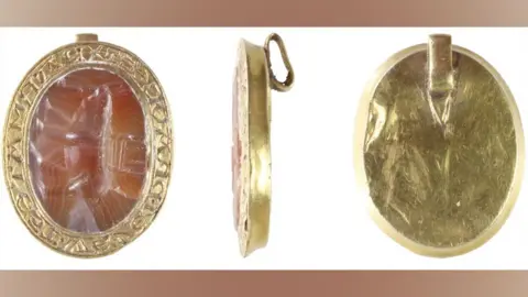 Andrew Williams/Norfolk County Council Three views of an oval medieval seal matrix, made from gold and a semi-precious gemstone. The view on the left shows its front. Its thin gold rim is etched with carved letters and surrounds the oval pink-toned gemstone, which is carved with an elephant with a castle or howdah on its back. The view in the middle is side on, showing a gold rim and a loop on its back. The view on the right shows plain gold back with a narrow gold rim and the loop at the top side. 