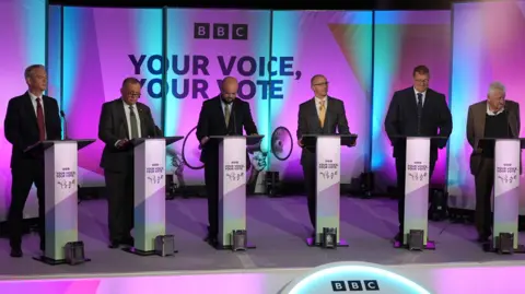 BBC All six candidates in the debate on stage standing at podiums with BBC Your Voice Your Vote branding behind them.