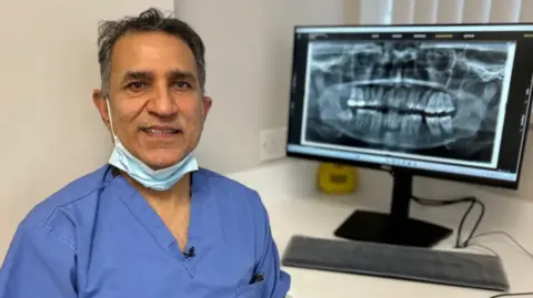 Dentist Shiv Pabary has a face mask under his chin and is smiling. He stands in front of a computer screen showing an X-ray of a mouth with teeth.