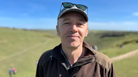 BBC/George Carden A man dressed in a brown top,  with a blue baseball cap with black sunglasses perched on it, is looking at the camera. Behind him is a green hillside and a blue sky