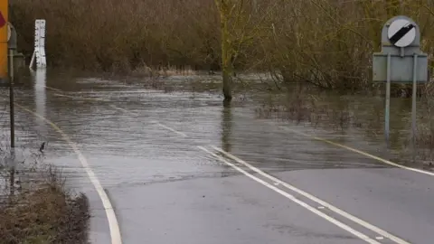 Shaun Whitmore/BBC A single carriageway tarmacked road disappearing under water. There are trees on the right sticking out of the water and an average speed sign. 