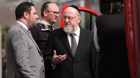 PA Media Chief Rabbi Sir Ephraim Mirvis gets out of a vehicle and speaks to a man in a grey suit