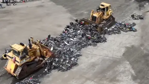 Two bulldozers scoop up the destroyed motorbikes and mopeds.
