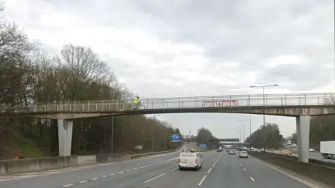 Google Three lanes of a motorway with a slip road to the left and a blue sign in the distance. There is a white van travelling ahead and cars in the distance too.