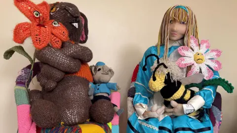A crafted brown rabbit holding an orange flower next to a crafted Alice holding a knitted bee. 
