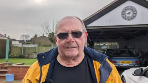 BBC A man in glasses, standing in front of a garage at his home, wears a black t-shirt and yellow coat