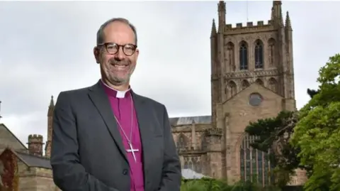 Bishop of Hereford Richard Jackson stands in front of the cathedral. He is wearing church attire and standing with his hands folded.