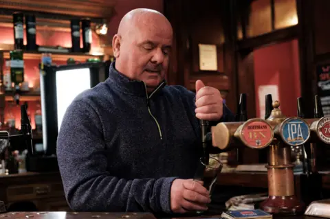 James Lacey pulling a pint on The Tall Ship set. He is wearing a dark top and looking down at the pint glass he is pouring the beer into from a pump. There are various spirits in the background.