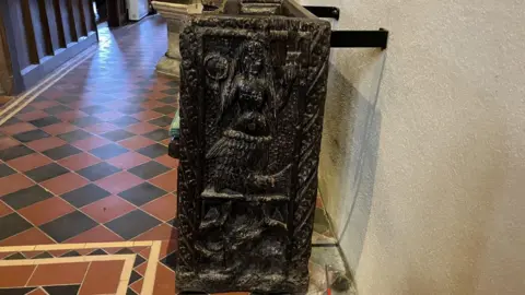 Etched onto a pew inside a church is the figure of a mermaid with long hair and carrying a comb. It sits on a floor of terracotta and black square tiles. 