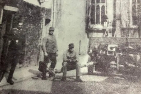 David Isherwood A black and white photo of German soldiers and guns from World War Two on a street corner. The men are all in uniform - one stands next to a doorway in an officer's uniform. Two dressed in ordinary combat uniforms are next to a large anti-tank gun, with one sitting on a part of it.