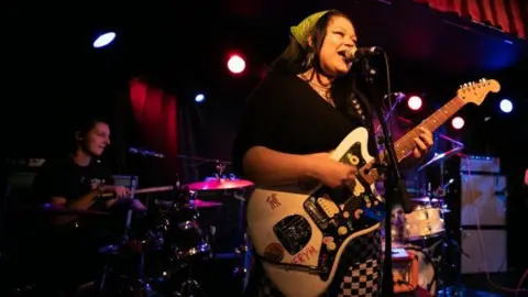 Getty Images Cherym se apresentando no palco em Dublin em 2023. Hannah canta em um microfone na frente do palco, segurando uma guitarra elétrica branca. Allanagh está atrás dela na bateria. 