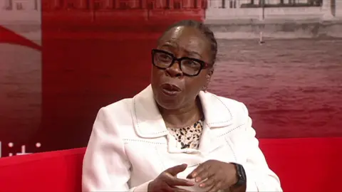 Paulette Hamilton in a white jacket over a pattered top. She is wearing heavy black framed glasses and is sitting in the TV studio, on a red sofa