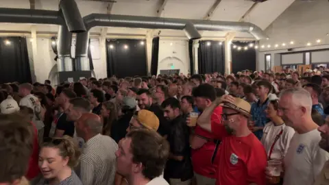 Fans are standing in rows watching a screen in a large menu.
