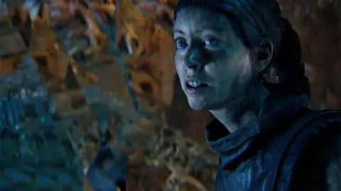 Ninja Theory A computer-generated, photo-realistic female character looks fearful as she spies something off-camera. She appears to be inside some sort of damp cave environment.