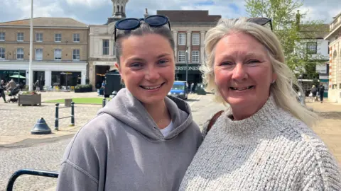 Holly Nichol/BBC Jan and Grace Moore standing in a market square in Hitchin. Grace is on the left, with pulled back mid brown hair, her dark glasses on the top of her head and wearing a grey hoodie. Jan is on the right and has blonde shoulder length hair, has her dark glasses on the top of her head and is wearing a beige funnel neck jumper. They are both smiling. 