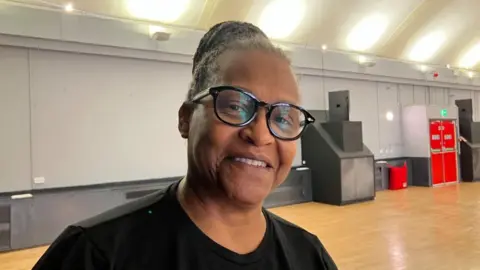 Mature woman, in a t-shirt, smiling at the camera, stood in a empty event space