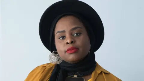 Muneera Pilgrim Muneera with a black hat and headwrap, wearing red lipstick, smiling at the camera