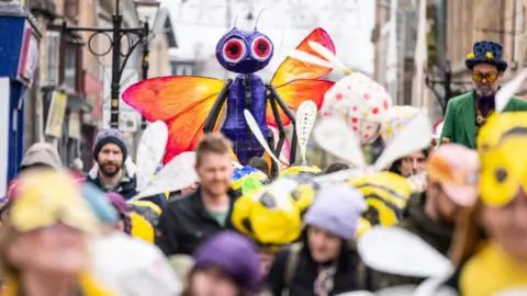 Paul Campbell A purple insect puppet with big eyes and colourful wings is held aloft above a crowd of people.