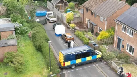 PA Media An aerial shot of the scene of a house explosion in Bristol. There are two red-brick properties, one of which has a boarded up downstairs window. Police and forensics officers and a police van can be seen in the street behind a cordon. There is also a forensics tent at the scene.