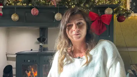 BBC Agnieszka McIntyre has shoulder-length brown hair and is wearing a white knitted jumper. She sits in front of a black metal wood-burning stove below a black fireplace decorated with Christmas baubles and red Christmas bows.