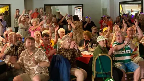 A large group of people dancing- some wear wigs and Glasgow Celtic green and white kits