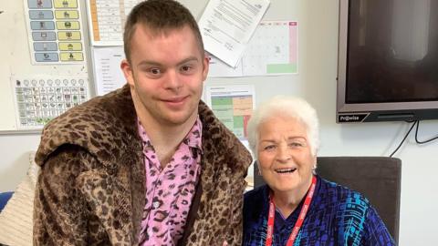 Smiling Jack and Pam St Clement posing for the camera. She is sitting on a chair and wears a blue top. He is standing next to her and wears a pink leopard top underneath a brown leopard coat. There is a TV and school posters behind them