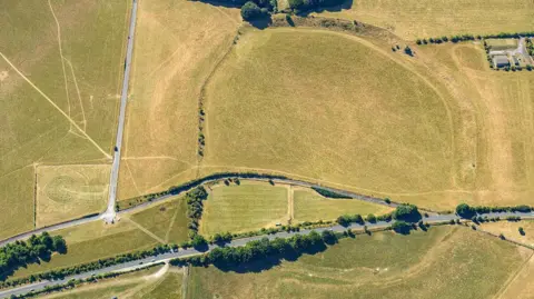 Historic England Archive/Heritage Images via Getty Images A drone shot of a green landscape with some trees at the bottom surrounding a road. The fields have lines in them, indicating paths and also ancient features